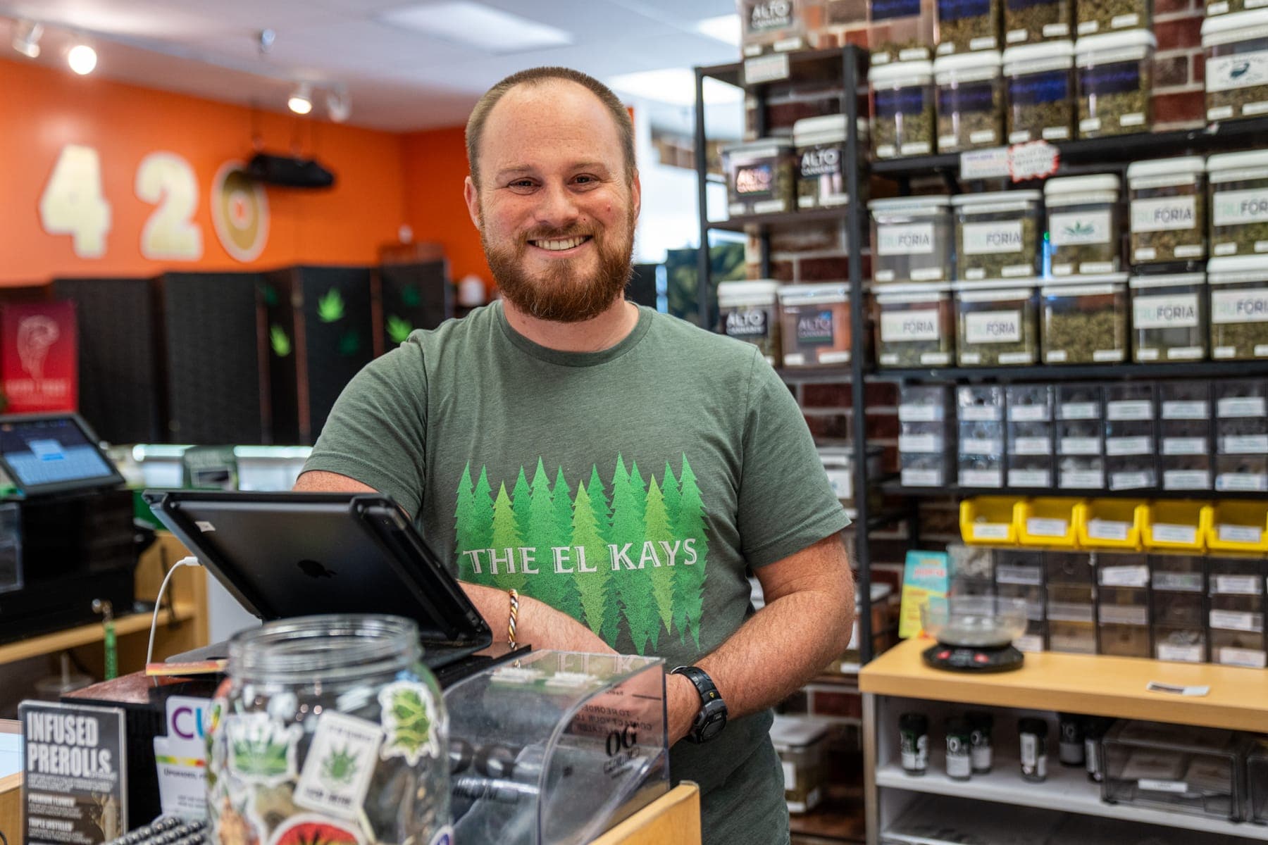 Stephen, one of our budtenders, at the register with the wall of flower jars behind him
