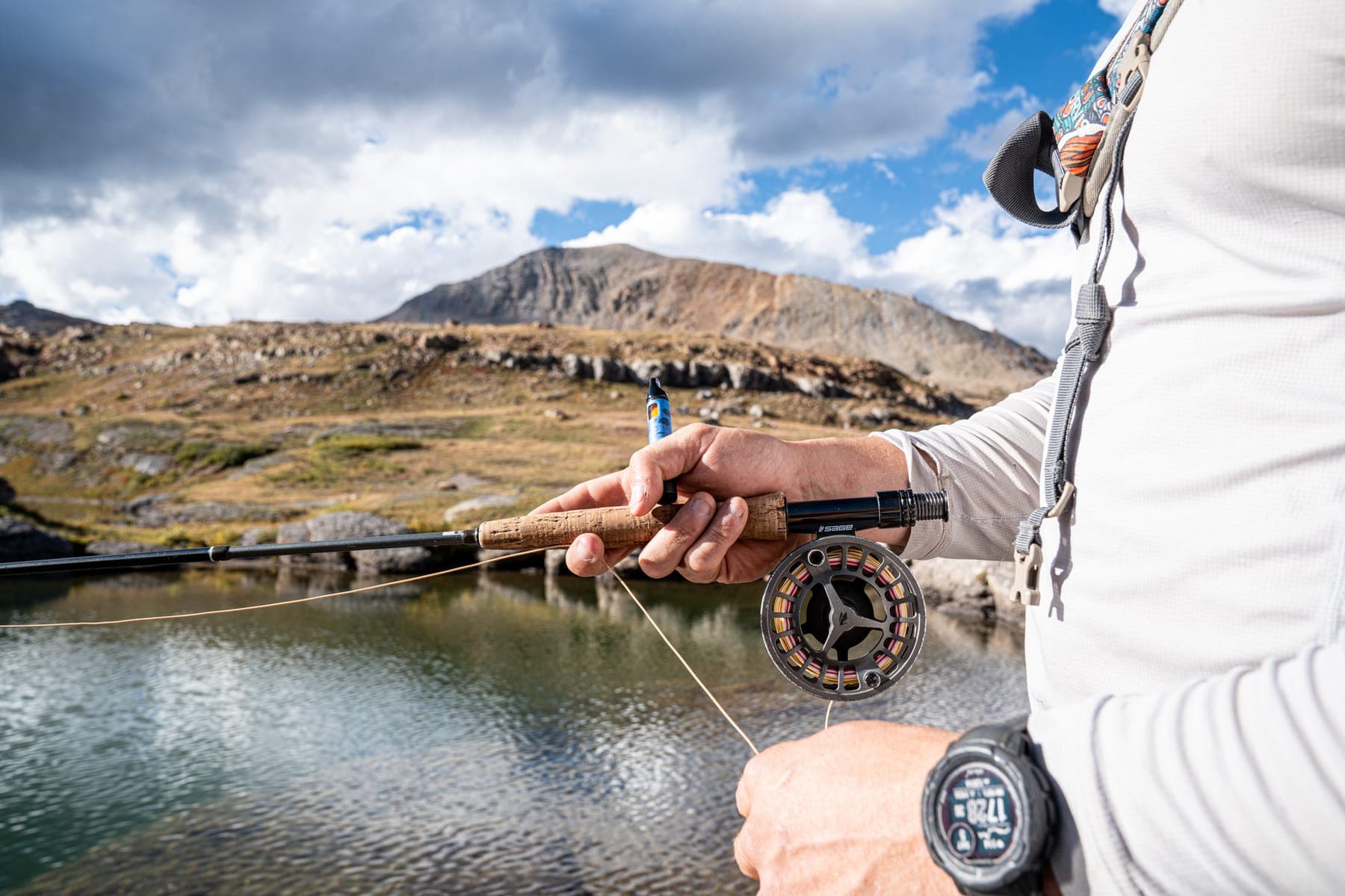 An angler working a fly reel at the edge of a high mountain lake under a big sky