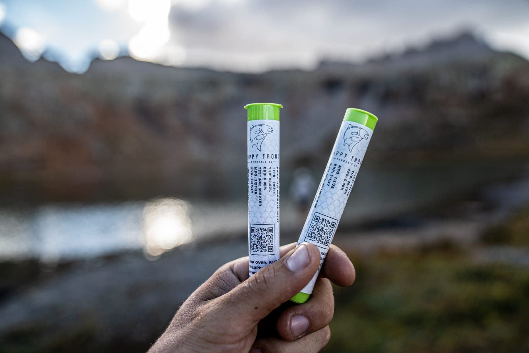 A hand holding two sealed pre-roll tubes above an alpine lake at dusk