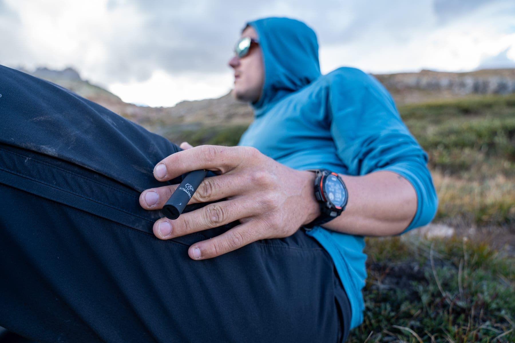 A hiker in a blue hoodie resting in the grass below a mountain ridge, a vape pen in hand