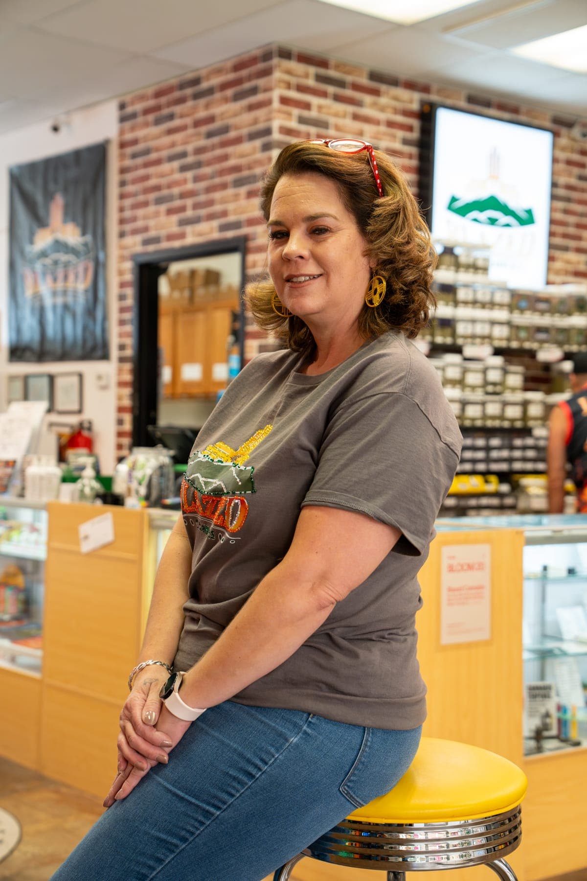 Jennifer, one of the owners, sitting on a yellow stool at the counter, the shop behind her