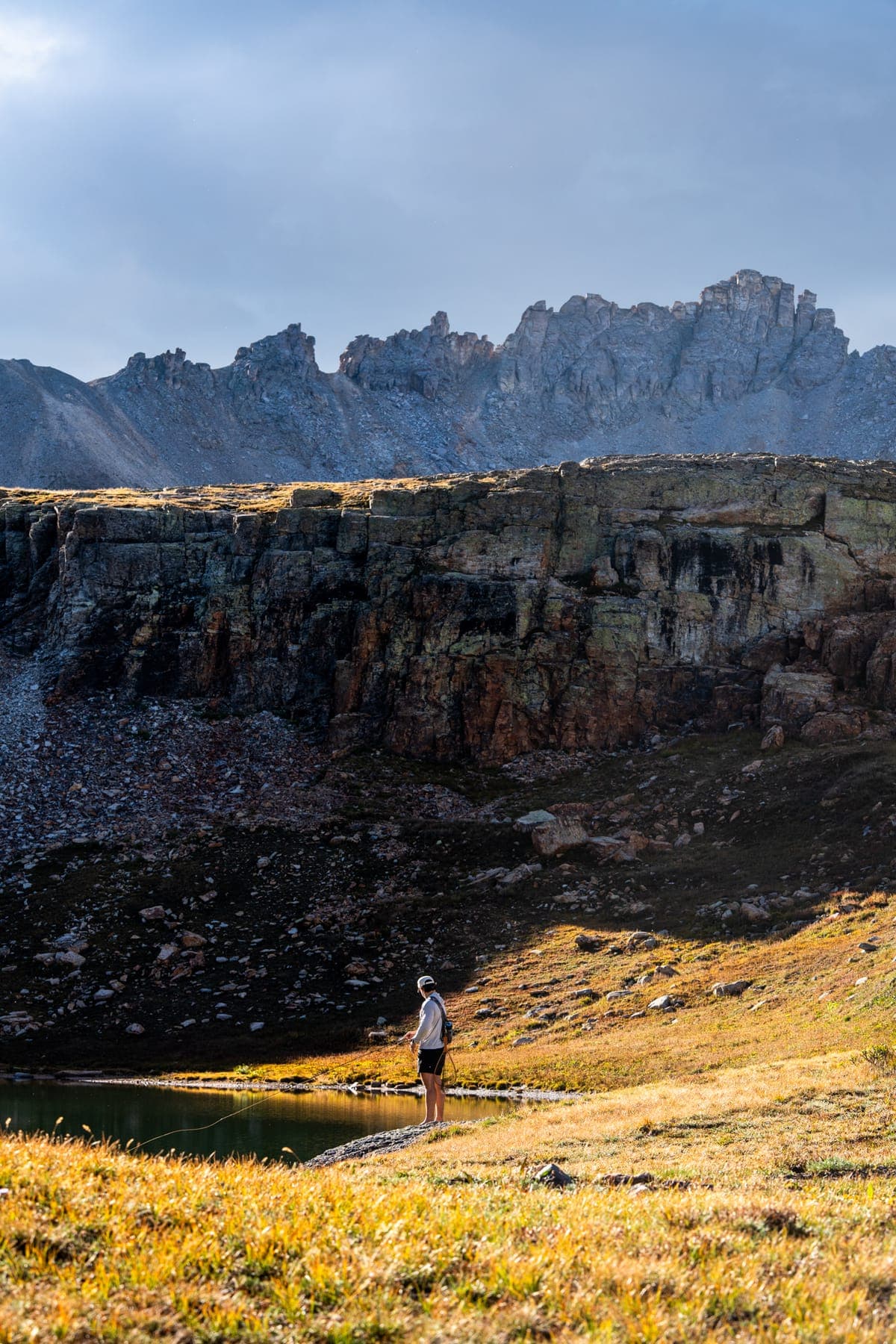 A lone angler on the shore of an alpine lake below jagged peaks, late light on the grass